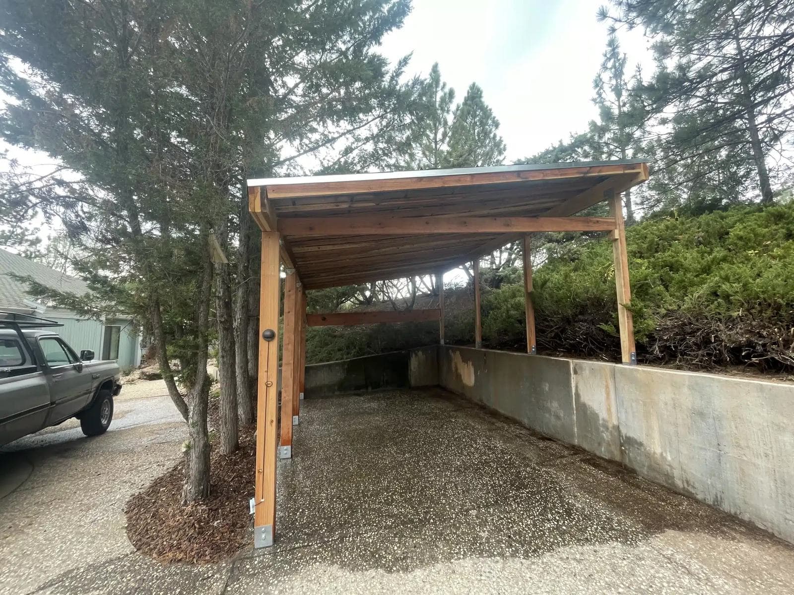 Timber-frame carport with a single-slope roof and integrated lighting, built against a concrete retaining wall on a hillside
